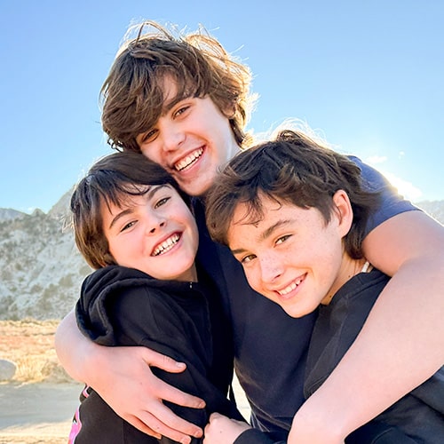 Three smiling brothers hugging outdoors, showcasing bright, healthy, and beautifully aligned smiles against a sunny sky and mountain backdrop.