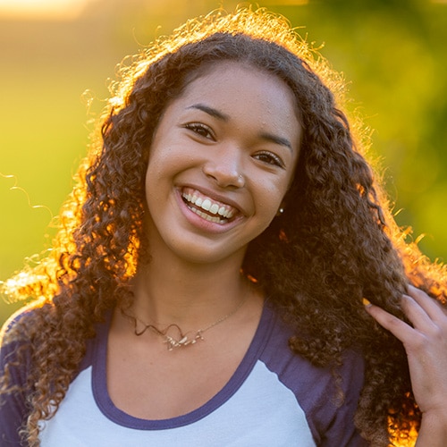 A young woman with long, curly hair smiling brightly outdoors, highlighting a beautifully straight and confident smile illuminated by warm golden hour sunlight.
