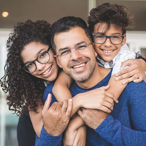 A happy father posing closely with his teenage daughter and son, all wearing glasses and smiling brightly to display healthy, confident, and straight teeth. Smithtown Orthodontics in Smithtown, NY