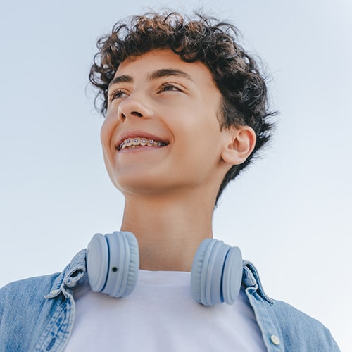 A teenage boy with curly brown hair smiling confidently while wearing traditional metal dental braces. He is looking upward and slightly to the side, with light blue headphones resting around his neck, a white t-shirt, and a light denim button-down shirt against a clear sky background.