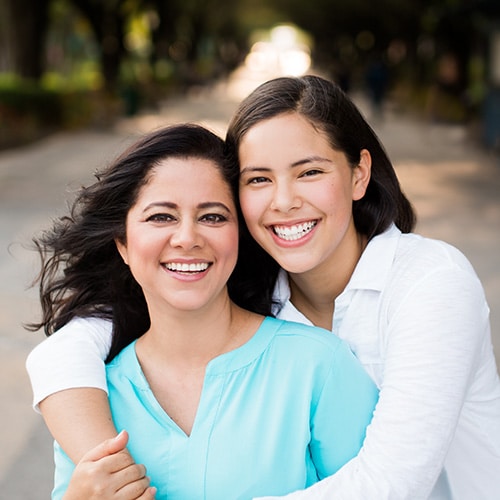 A mother and her teenage daughter smiling warmly while posing together outdoors, showcasing two generations of bright, healthy, and beautifully straight smiles.