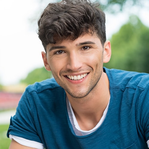 A smiling young man with dark, textured hair wearing a teal t-shirt outdoors, highlighting a bright, healthy, and perfectly straight smile. Smithtown Orthodontics in Smithtown, NY