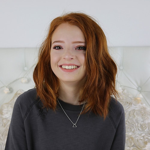 A young woman with shoulder-length, wavy red hair smiling brightly to show off a straight, healthy smile. She is wearing a dark grey sweatshirt and a simple silver triangle necklace against a soft, neutral background.