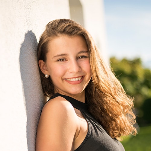 A teenage girl leaning against a white textured wall outdoors, smiling warmly to showcase a straight, confident smile. She has long, wavy light brown hair, is wearing a black sleeveless top, and stands under bright sunlight with a blurred green background.