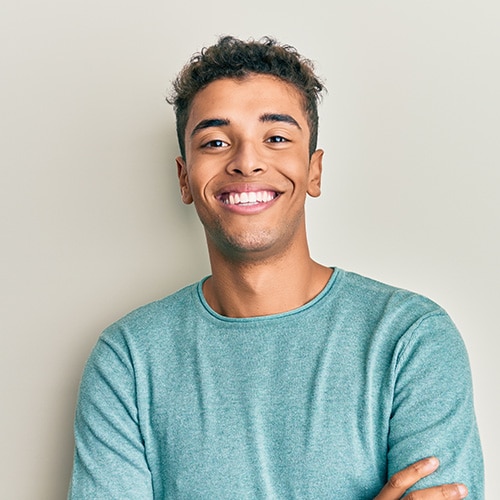 A young man with short, curly hair wearing a mint green long-sleeve shirt smiling confidently with arms crossed, showcasing a bright and perfectly aligned smile. Bienvenidos a Smithtown Orthodontics en Smithtown, NY