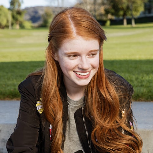 Smiling young woman with long red hair sitting outdoors in a park, representing a confident and healthy smile for orthodontic care content.
