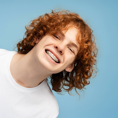 A smiling teenage boy with curly red hair leaning forward against a light blue background, proudly showing off his orthodontic braces.